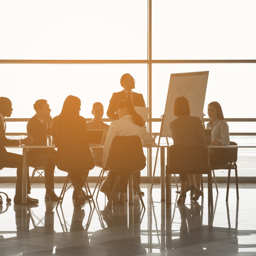 Group of people sitting at the office table while their partner standing near board with chart. Panoramic window on background