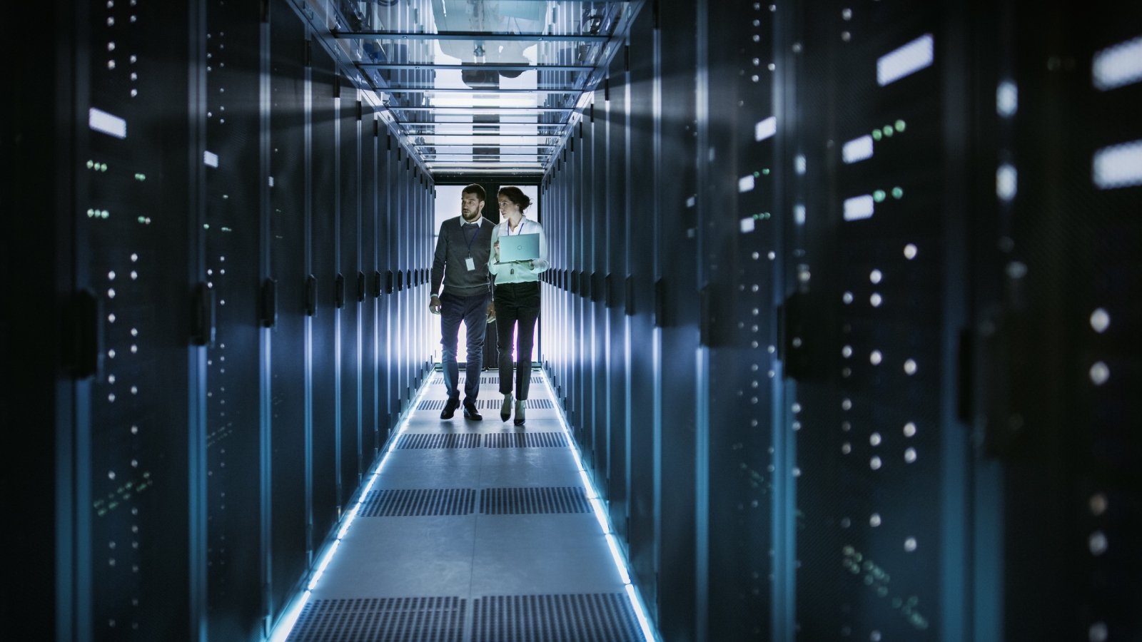 A man and a women walking down a corridor surrounded by racks in a data centre