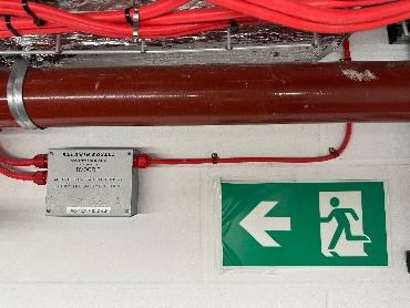 an image showing red pipes and cables on a wall and an emergency exit sign
